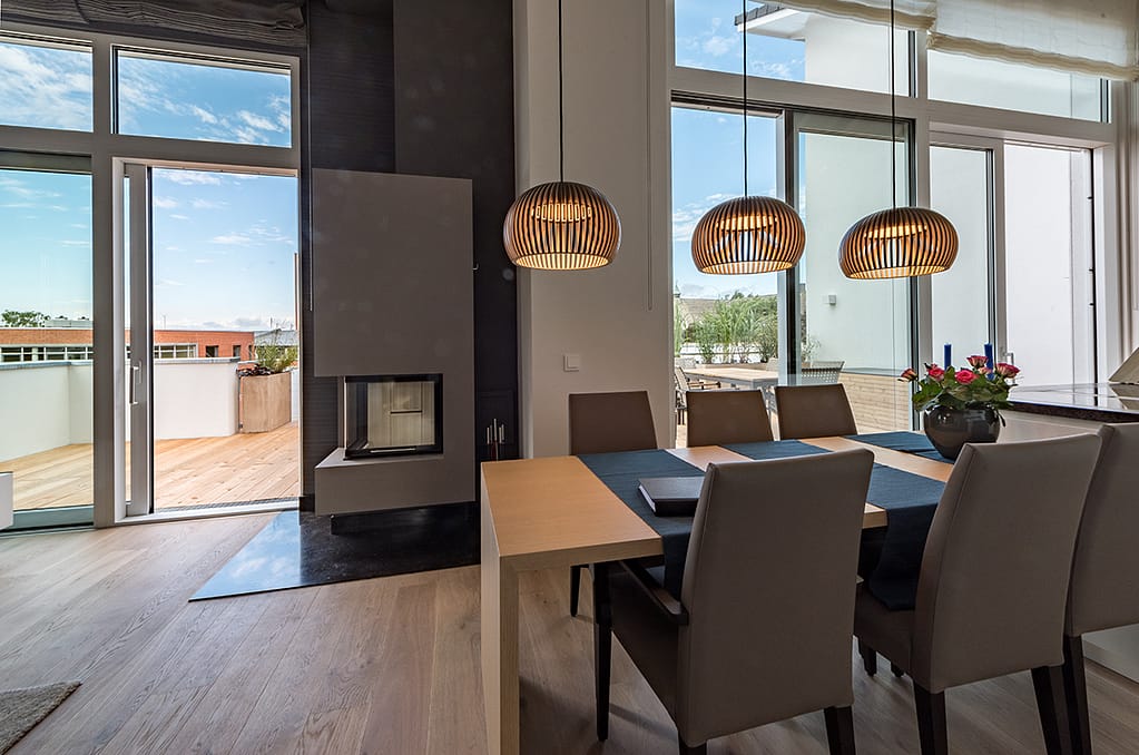 Moderner Essbereich mit rechteckigem Holztisch, sechs gepolsterten Stühlen, drei hängenden Holzlampen, großem Fenster mit Blick auf Terrasse und Kamin mit schwarzer Bodenplatte.
