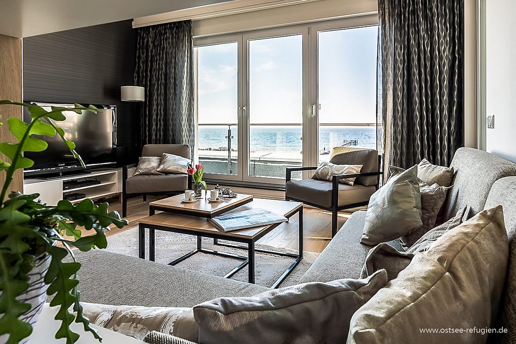 Modern eingerichtetes Wohnzimmer mit grauem Sofa, Sesseln, einem Couchtisch mit Tee-Set und Vase mit roten Tulpen vor großen Fenstern mit Blick auf das Meer.