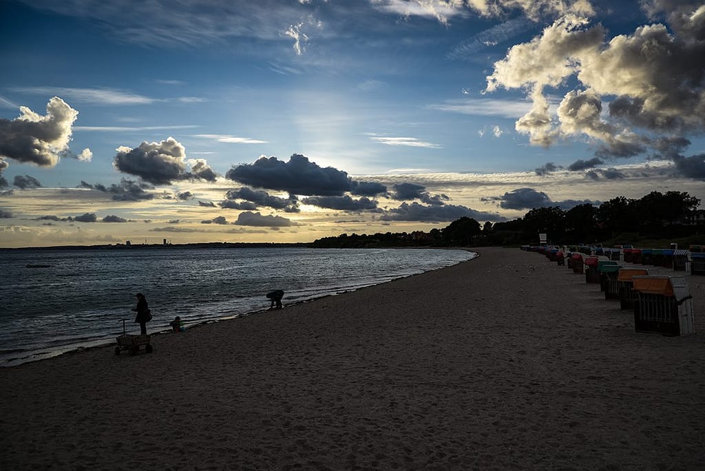 Düsterer Strand mit drei Personen am Wasser, Reihe von bunten Strandkörben rechts, dramatischer Himmel mit Wolken bei Sonnenuntergang