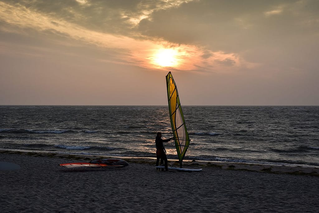 Strand bei Sonnenuntergang mit einer Person, die ein Windsurfbrett mit einem gelb-grünen Segel hält, während das Meer im Hintergrund ruhig ist und weitere Windsurf-Ausrüstung auf dem Sand liegt.