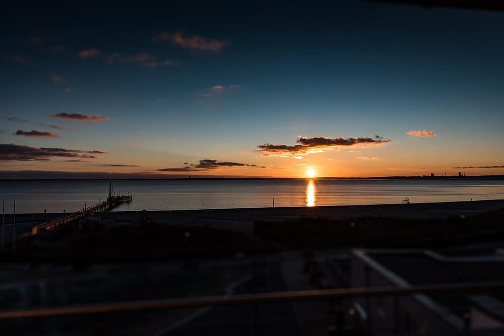 Sonnenuntergang über ruhigem Meer mit langem Steg, der ins Wasser führt, und wenigen Wolken am Himmel