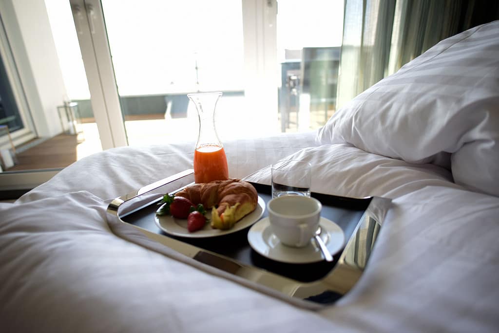 Frühstückstablett auf weißem Bett mit Croissant, Erdbeeren, einem Glas Orangensaft, einer Kaffeetasse und einem leeren Glas vor hellem Fenster und grünen Vorhängen.