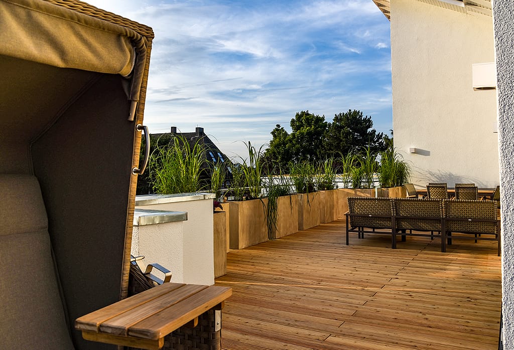 Balkon mit Holzfußboden, bepflanzten Blumenkästen entlang des Geländers, mehreren geflochtenen Gartenmöbeln und einem großen Strandkorb mit aufklappbarem Tisch im linken Vordergrund