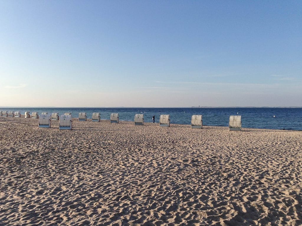 Leerer Sandstrand mit mehreren weißen Strandkörben in einer Reihe nahe am Wasser unter blauem Himmel