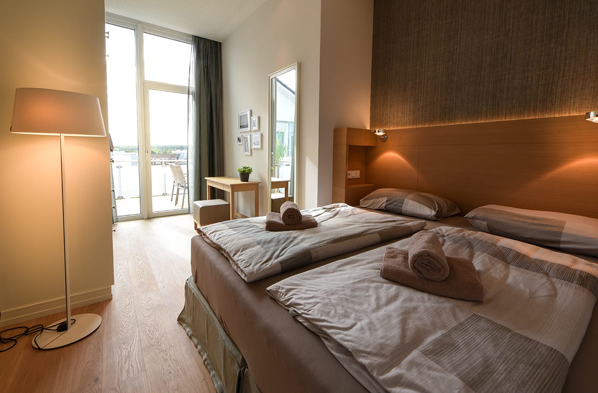 Modernes Schlafzimmer mit zwei Einzelbetten, jeweils mit gerollten braunen Handtüchern und Tagesdecken, stehendem Lampenlicht, Holzboden, Tisch mit Hocker und großem Fenster mit Blick auf Balkon und Außenterrasse.