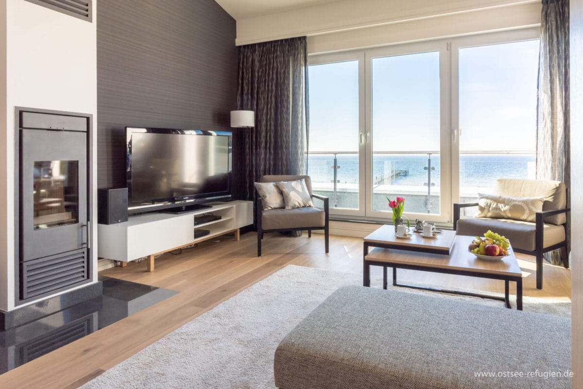 Helles Wohnzimmer mit großen Fenstern und Blick auf das Meer, zwei graue Sessel mit Kissen, moderner Kamin, Flachbildfernseher auf einem weißen Lowboard, ein Couchtisch mit Kuchen und Obst, helle Holzböden und ein graues Sofa am Rand eines beigen Teppichs