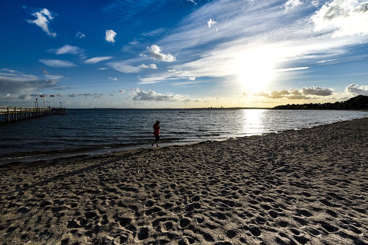 ein Strand mit vielen Fußabdrücken im Sand, eine Person in rotem Hemd am Ufer eines ruhigen Meeres mit einem Pier links im Hintergrund und einem hellen Sonnenuntergang am Himmel mit verstreuten Wolken
