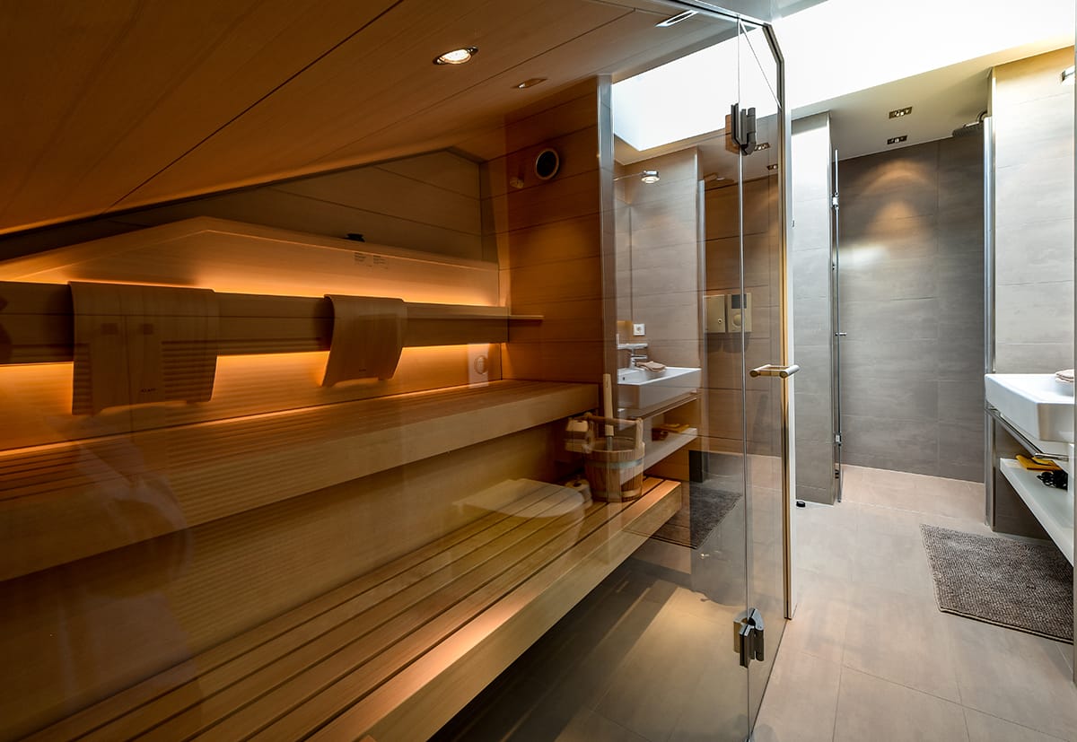Moderne Sauna mit Holzinnenverkleidung, integraler Holzbänke und warmem indirektem Licht, angrenzendes Badezimmer mit grauen Fliesen, Glasduschtür und weißen Waschbecken.