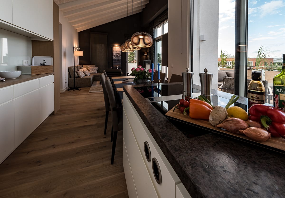 Moderner offener Wohnraum mit Kücheninsel, Esstisch und angeschlossenem Wohnzimmer, mit Holzfußboden und großen Fenstern, frischem Gemüse und Gewürzen auf der Kücheninsel und Blick auf eine Terrasse im Hintergrund.