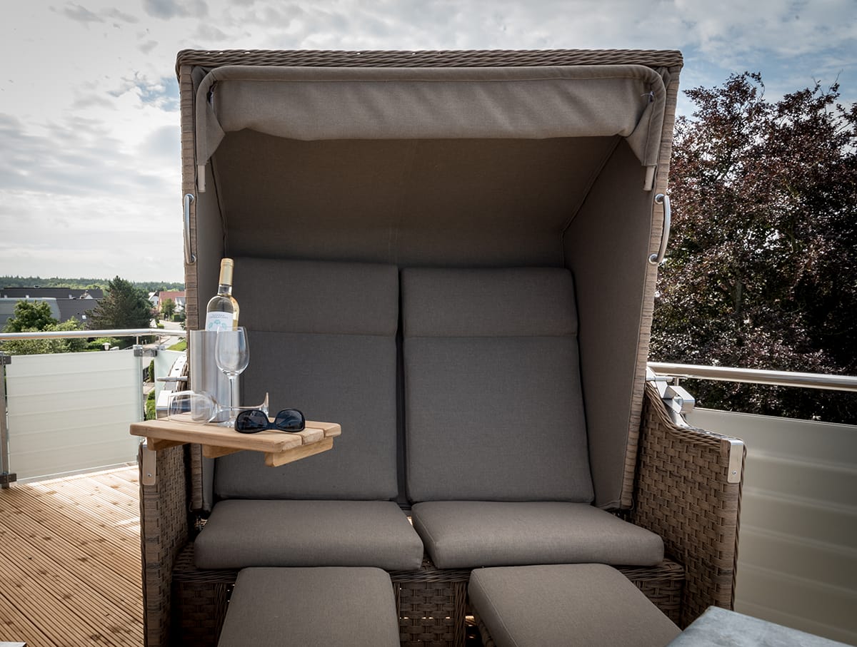 Ein geflochtener Strandkorb mit grauen Kissen auf einem Balkon, daneben ein kleiner Holzklapptisch mit einer Flasche Wein, zwei Weingläsern und einer Sonnenbrille, im Hintergrund Bäume und Himmel mit Wolken.