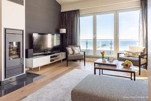 Helles Wohnzimmer mit großen Fenstern und Blick auf das Meer, zwei graue Sessel mit Kissen, moderner Kamin, Flachbildfernseher auf einem weißen Lowboard, ein Couchtisch mit Kuchen und Obst, helle Holzböden und ein graues Sofa am Rand eines beigen Teppichs