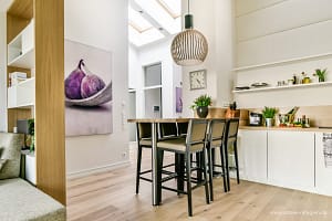 Moderner Küchenbereich mit Holzboden, einer hohen Tafel mit vier schwarzen Stühlen, einer großen hängenden Lampe, weißer Wand mit einem Bild von Feigen, einer Küchenzeile mit Pflanzen, Gewürzen und Küchenutensilien sowie einem großen Fenster im Dach für natürliches Licht.