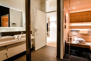 Moderner Badezimmerbereich mit Doppelwaschbecken, beleuchtetem Spiegel und offener Tür zum angrenzenden Raum, daneben sichtbare Holzwand und Glasabtrennung einer Sauna mit gefalteten Handtüchern und Sauna-Accessoires.