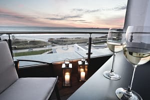 Balkon mit zwei Weingläsern auf einem Tisch, einer Polstersessel und zwei Laternen mit Kerzen, Blick auf das Meer bei Sonnenuntergang mit rosa Wolken am Himmel.