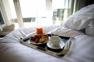 Frühstückstablett auf weißem Bett mit Croissant, Erdbeeren, einem Glas Orangensaft, einer Kaffeetasse und einem leeren Glas vor hellem Fenster und grünen Vorhängen.