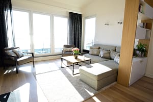 Hell beleuchtetes Wohnzimmer mit großer Fensterfront, grauem Ecksofa mit Kissen, zwei Sesseln, einem quadratischen Couchtisch mit Blumenvasen und hellbeigem Teppich auf Holzboden, daneben ein offenes Regal mit Pflanzen und Büchern.