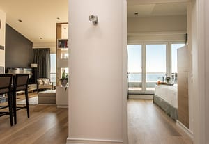 Blick durch eine zweiseitige Wand in eine modern eingerichtete Wohnung mit Holzboden, links Wohnbereich mit Stühlen, Sessel, Vorhängen und Fenster mit Meerblick, rechts Schlafzimmer mit Bett, einer Frühstückstablett und großen Fenstern mit Blick auf das Meer