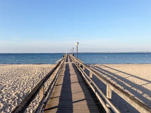 Holzsteg mit Laternen am Strand, der geradewegs aufs ruhige Meer unter blauem Himmel führt, mit Sand auf beiden Seiten des Stegs.