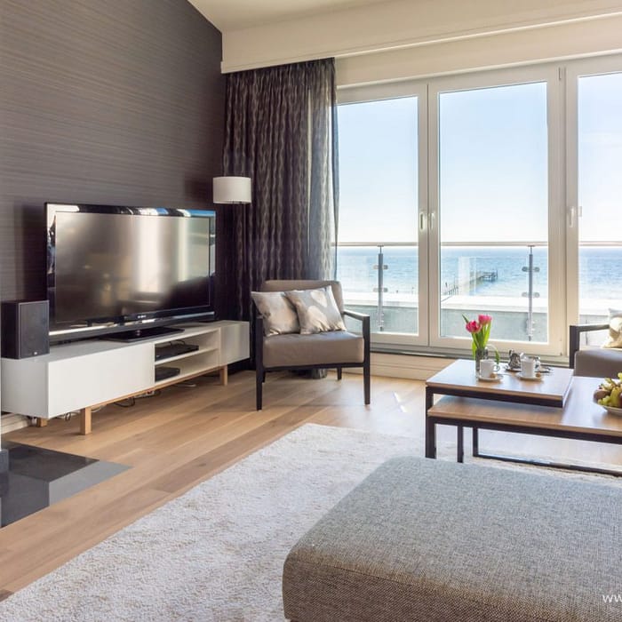 Helles Wohnzimmer mit großen Fenstern und Blick auf das Meer, zwei graue Sessel mit Kissen, moderner Kamin, Flachbildfernseher auf einem weißen Lowboard, ein Couchtisch mit Kuchen und Obst, helle Holzböden und ein graues Sofa am Rand eines beigen Teppichs