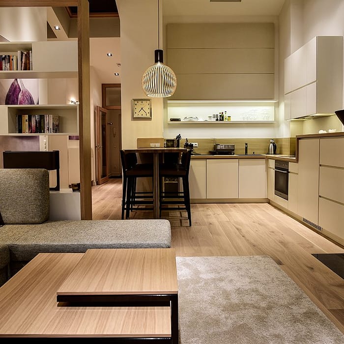 Modern eingerichtetes Wohnzimmer mit grauem Ecksofa, Holztisch, offenem Bücherregal und angrenzender offener Küche mit weißen Schränken und Holzboden, in warmer Beleuchtung.
