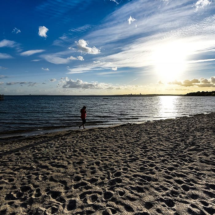 ein Strand mit vielen Fußabdrücken im Sand, eine Person in rotem Hemd am Ufer eines ruhigen Meeres mit einem Pier links im Hintergrund und einem hellen Sonnenuntergang am Himmel mit verstreuten Wolken
