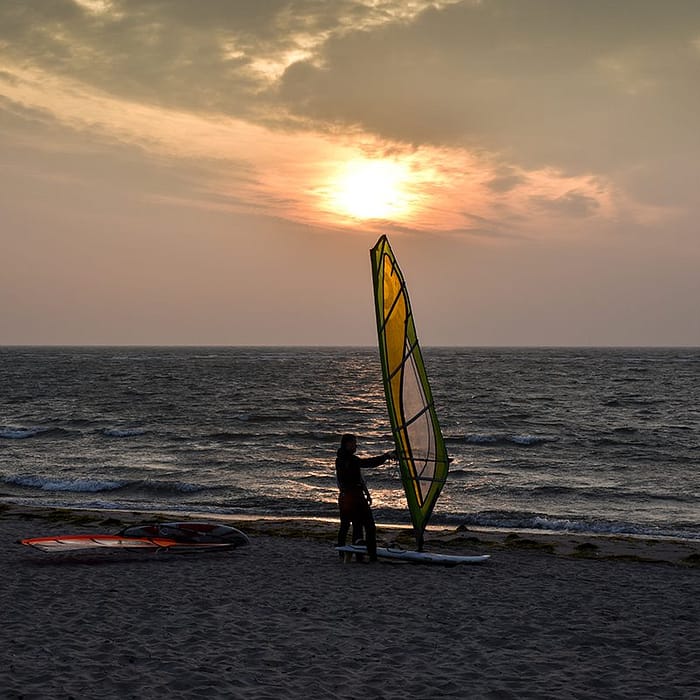 Strand bei Sonnenuntergang mit einer Person, die ein Windsurfbrett mit einem gelb-grünen Segel hält, während das Meer im Hintergrund ruhig ist und weitere Windsurf-Ausrüstung auf dem Sand liegt.