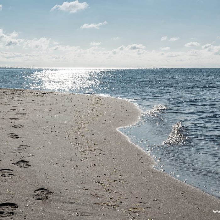 Küstenlinie mit Sandstrand und Fußspuren, im Hintergrund das ruhige Meer unter einem leicht bewölkten Himmel mit Sonnenreflexion auf dem Wasser.