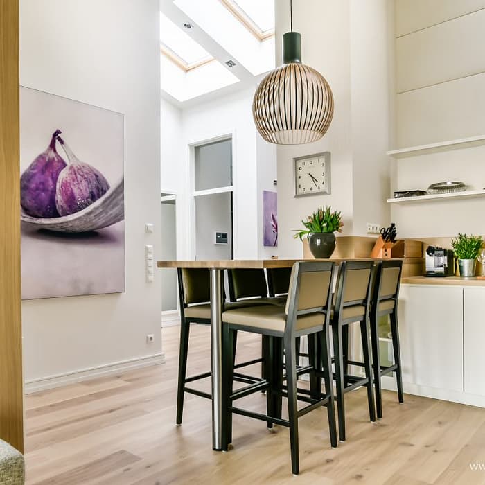 Moderner Küchenbereich mit Holzboden, einer hohen Tafel mit vier schwarzen Stühlen, einer großen hängenden Lampe, weißer Wand mit einem Bild von Feigen, einer Küchenzeile mit Pflanzen, Gewürzen und Küchenutensilien sowie einem großen Fenster im Dach für natürliches Licht.