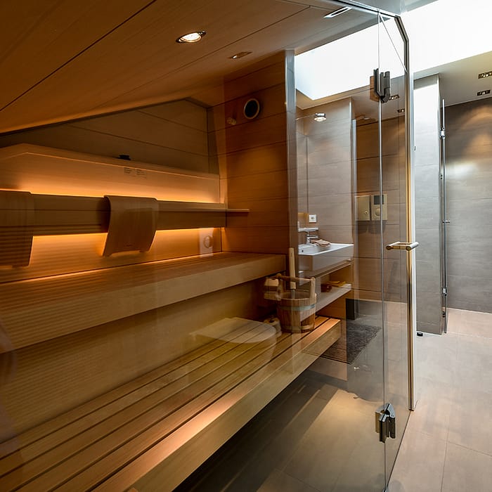 Moderne Sauna mit Holzinnenverkleidung, integraler Holzbänke und warmem indirektem Licht, angrenzendes Badezimmer mit grauen Fliesen, Glasduschtür und weißen Waschbecken.
