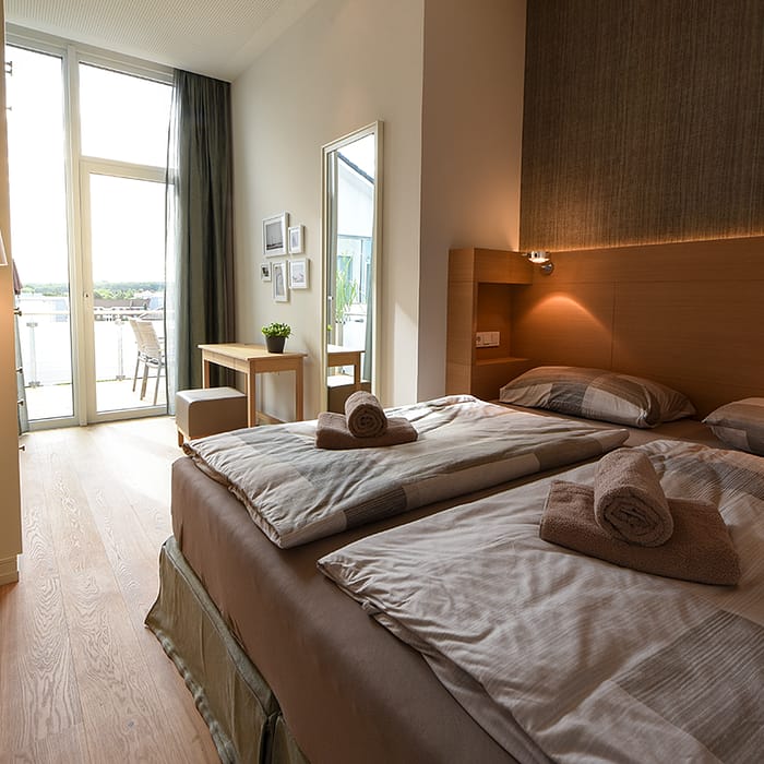 Modernes Schlafzimmer mit zwei Einzelbetten, jeweils mit gerollten braunen Handtüchern und Tagesdecken, stehendem Lampenlicht, Holzboden, Tisch mit Hocker und großem Fenster mit Blick auf Balkon und Außenterrasse.