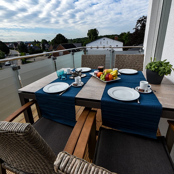 Balkon mit Tisch, der mit blauen Läufern, weißem Geschirr, Tassen, Besteck und einer Platte mit Obst sowie einem Blumentopf dekoriert ist, umgeben von Stühlen mit geflochtenen Rückenlehnen und dunklen Sitzkissen, Blick auf eine Nachbarschaft mit Häusern und bewölktem Himmel.