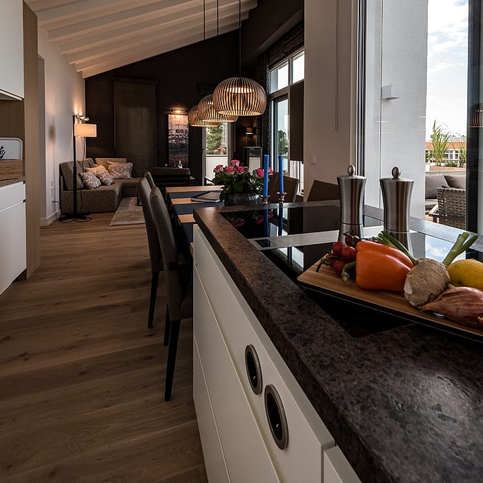Moderner offener Wohnraum mit Kücheninsel, Esstisch und angeschlossenem Wohnzimmer, mit Holzfußboden und großen Fenstern, frischem Gemüse und Gewürzen auf der Kücheninsel und Blick auf eine Terrasse im Hintergrund.