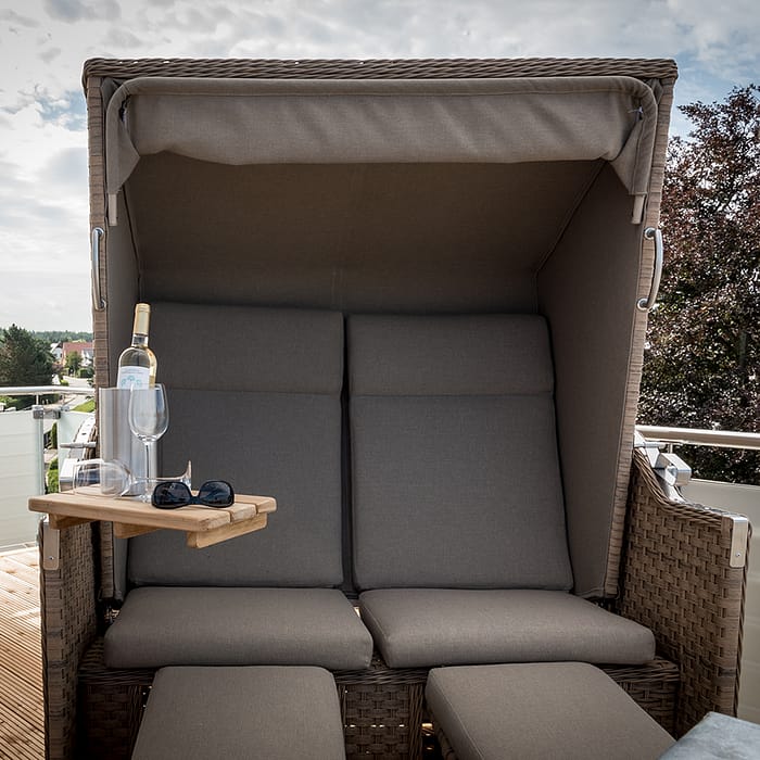 Ein geflochtener Strandkorb mit grauen Kissen auf einem Balkon, daneben ein kleiner Holzklapptisch mit einer Flasche Wein, zwei Weingläsern und einer Sonnenbrille, im Hintergrund Bäume und Himmel mit Wolken.